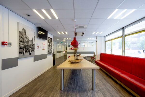 Moderne wachtruimte aan de Jan Tooropstraat met een lange tafel, rode bank en zwart-witfoto’s aan de muur.